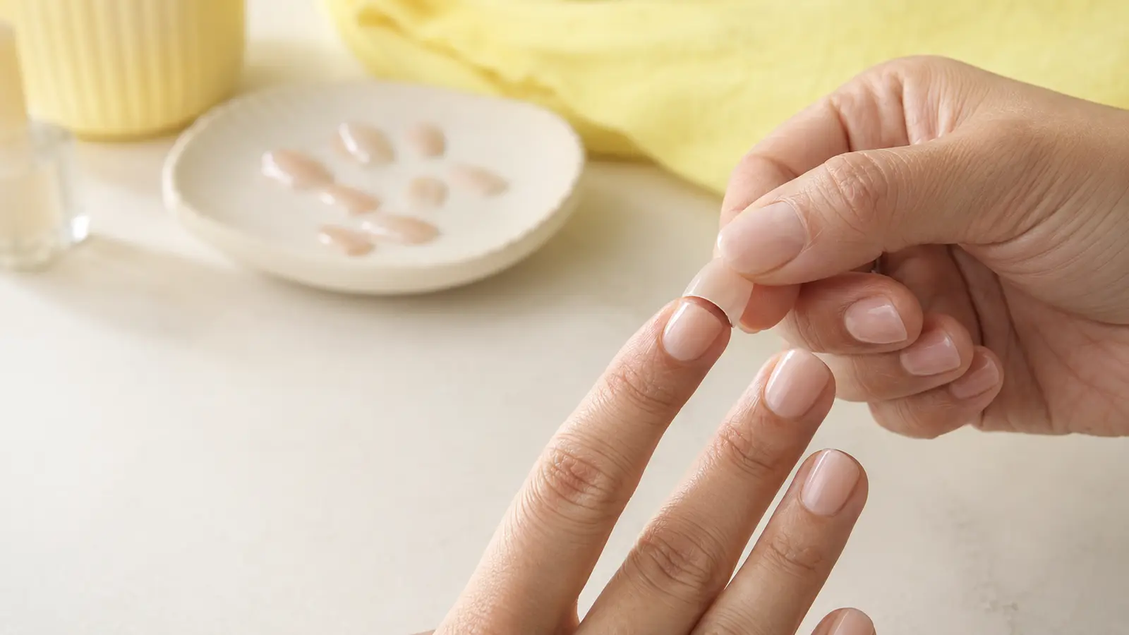 A loose nude press-on nail being gently lifted from an index finger at a clean home manicure desk.